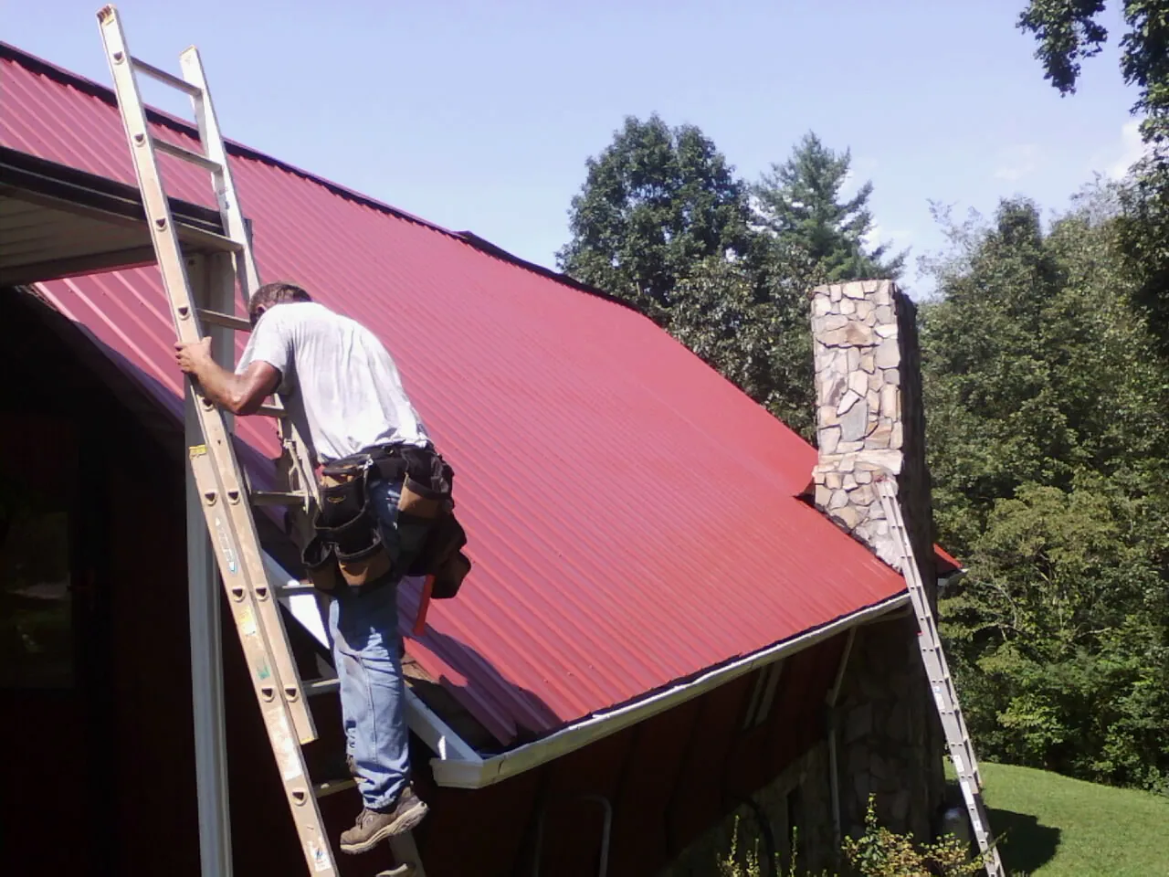 Roofer on a ladder beside a red metal roof and stone chimney — Soffit & Fascia Repair by Roofing Brothers in Taylorsville